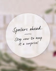 A blurred background of wrapped stationery gifts with a white circle overlay in the centre reading: “Spoilers ahead… Stop now to keep it a surprise!” in handwritten-style text. filter_alt_text