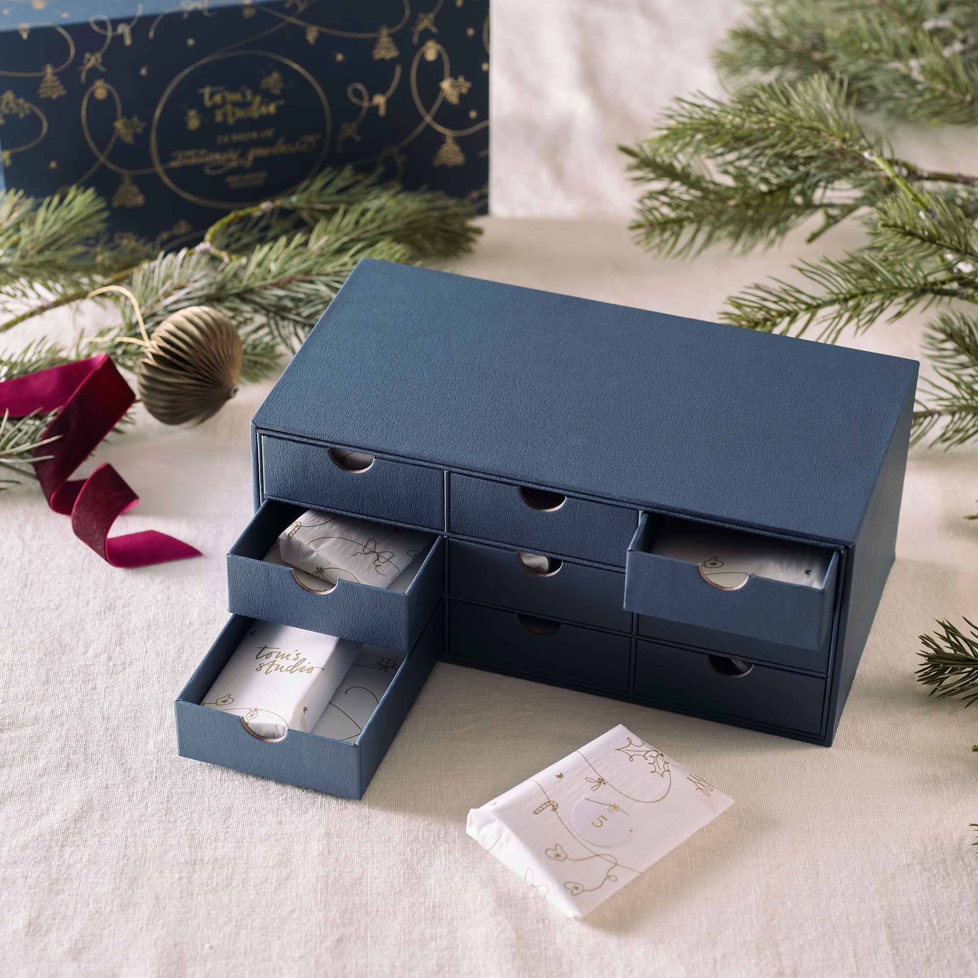 The Tom’s Studio Advent Calendar open, showing a navy drawer-style box with individually wrapped numbered gifts inside, surrounded by festive greenery.