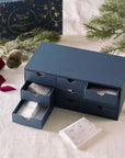 The Tom’s Studio Advent Calendar open, showing a navy drawer-style box with individually wrapped numbered gifts inside, surrounded by festive greenery.