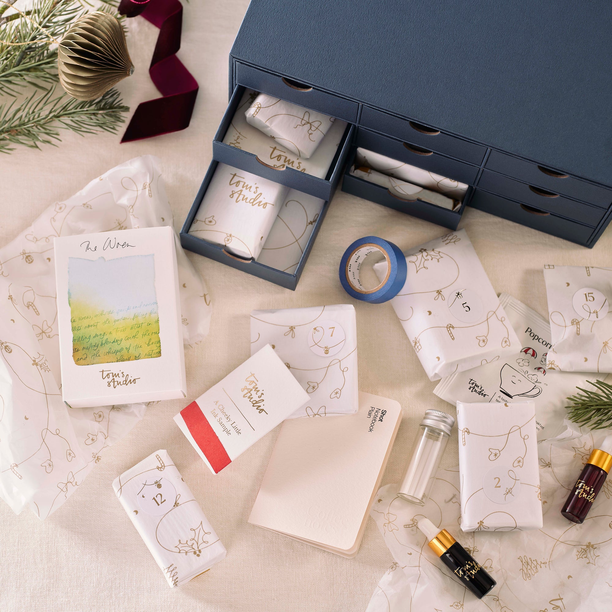 Contents of the Tom’s Studio Advent Calendar displayed on a table: wrapped gifts, mini ink bottles, washi tape, a notebook, and other stationery items, with the drawer box in the background. 
filter_alt_text