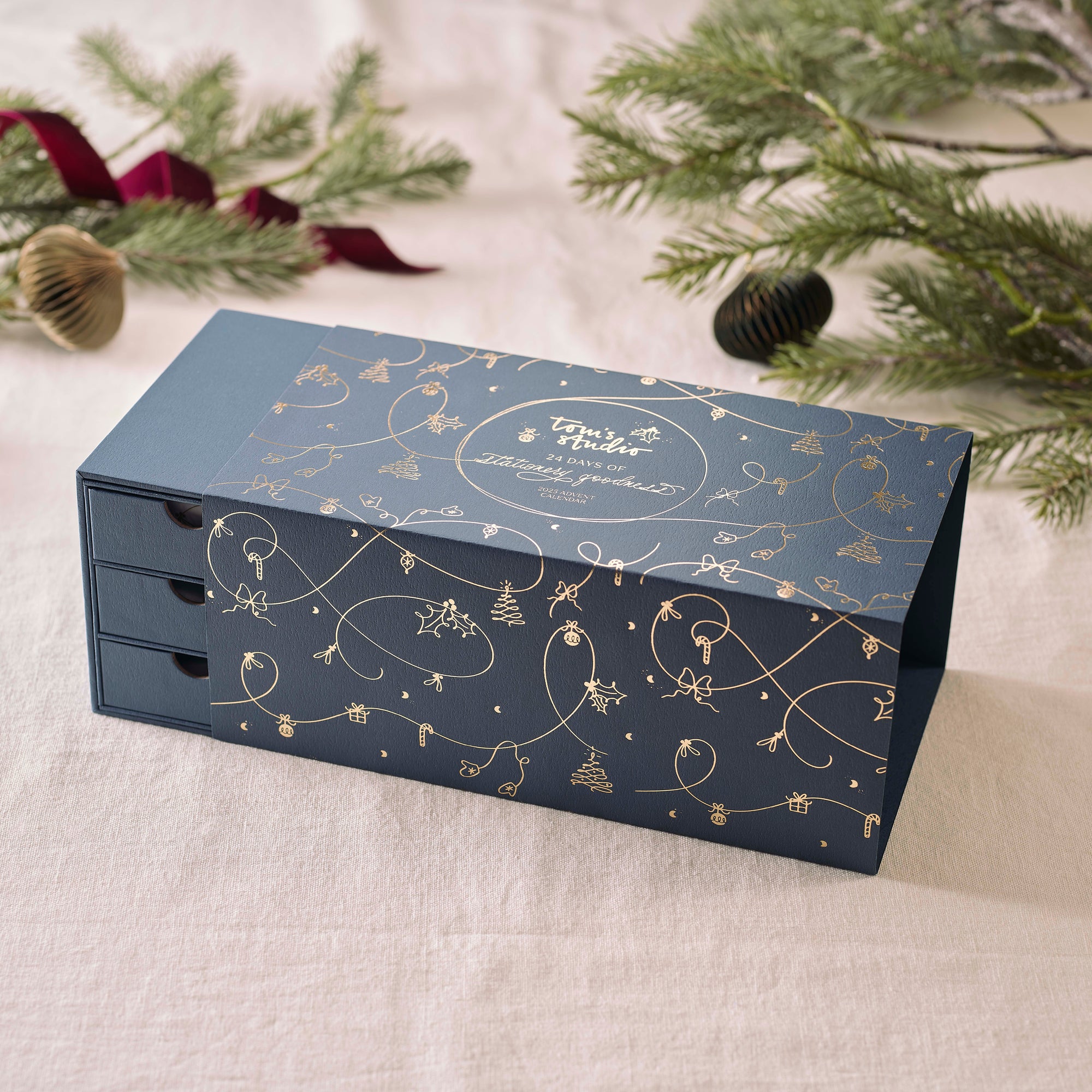 A navy blue Tom’s Studio advent calendar box with three drawers, decorated with delicate gold foil illustrations of Christmas motifs, placed on a light fabric with festive greenery.
