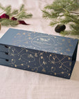 A navy blue Tom’s Studio advent calendar box with three drawers, decorated with delicate gold foil illustrations of Christmas motifs, placed on a light fabric with festive greenery.