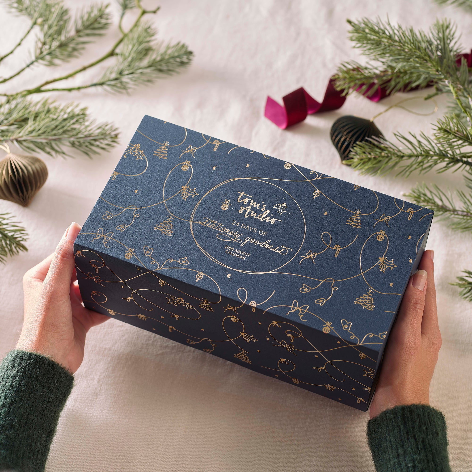 A pair of hands holding the closed Tom’s Studio advent calendar box, navy blue with gold foil illustrations of Christmas decorations, surrounded by pine branches and ribbon.