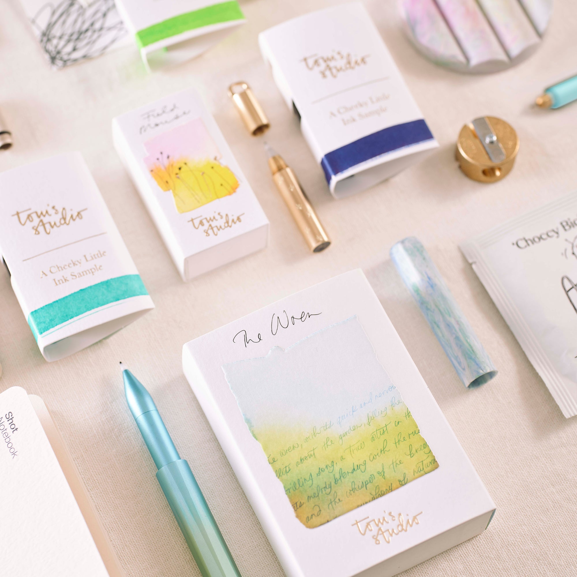 A close-up of unwrapped advent calendar gifts from Tom’s Studio, including mini ink samples, a pen, notebooks, and accessories, styled on a cream fabric background. filter_alt_text