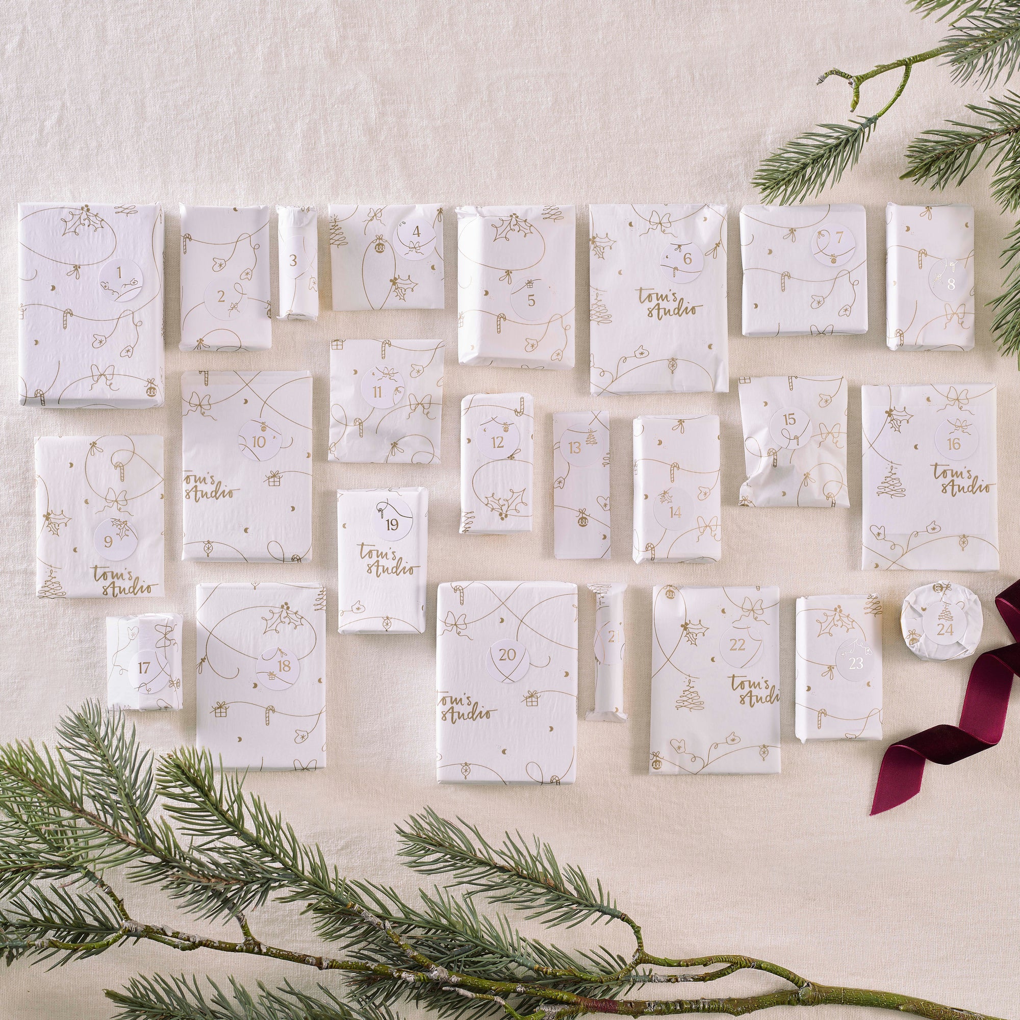 Twenty-four individually wrapped Tom’s Studio advent calendar gifts in white and gold wrapping paper, neatly arranged on a cream background with pine branches.