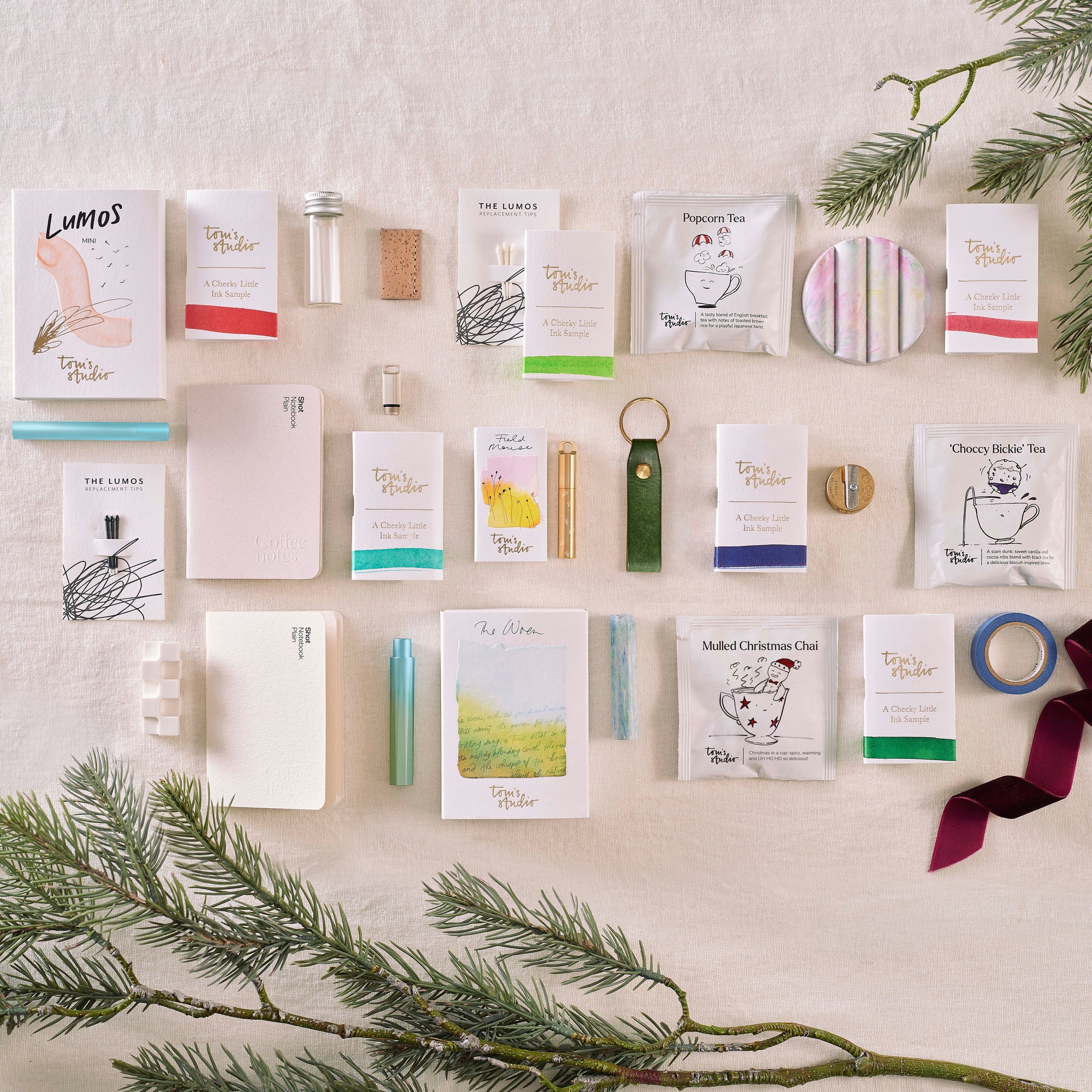 The full contents of the Tom’s Studio advent calendar laid out, including pens, ink samples, notebooks, tea sachets, and stationery accessories, styled with festive greenery and ribbon. filter_alt_text