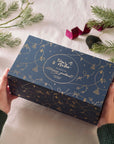Hands holding a navy and gold illustrated Tom’s Studio Advent Calendar box with festive decorations and pine branches in the background.