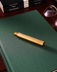 A Brass Wren refillable writing pen , capped, rests on top of a dark green notebook, placed beside glasses, a laptop, and stacked paperbacks on a wooden desk.