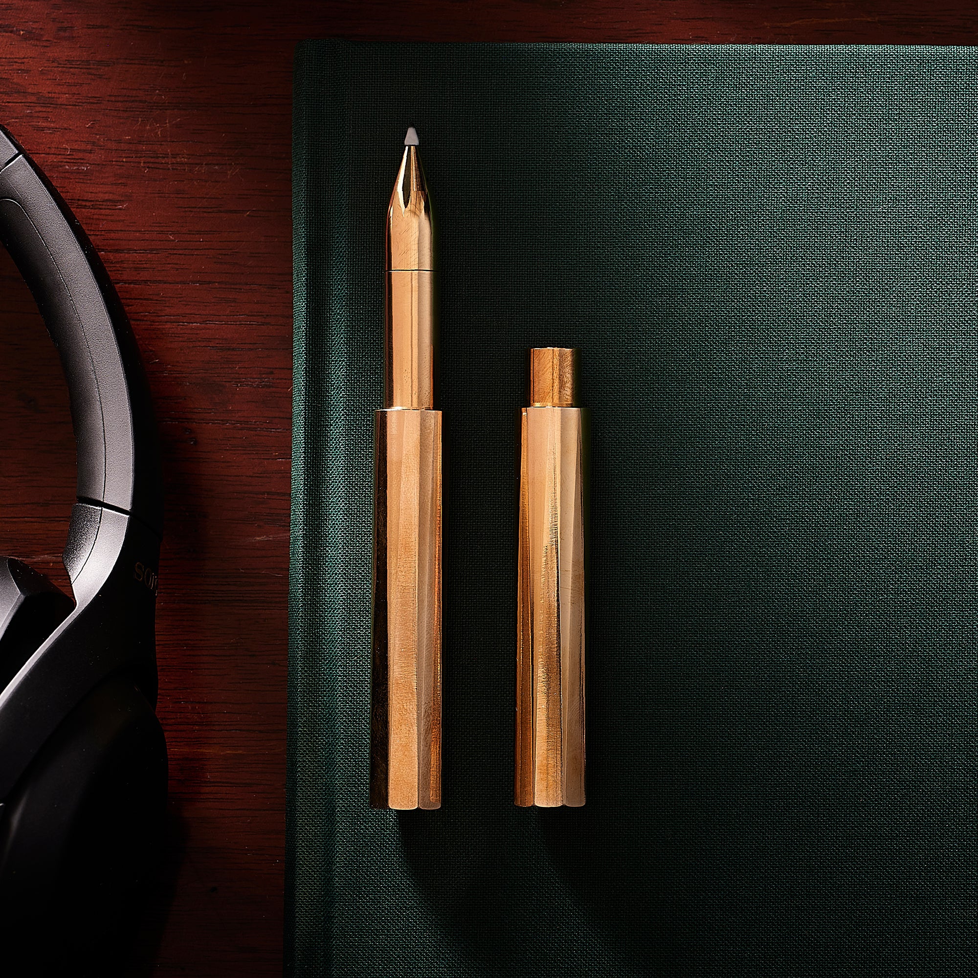 Two Brass Wren refillable writing pens – one capped and one with the lid posted – ontop of a green notebook, with glasses and headphones on the wooden desk nearby.