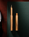 Two Brass Wren refillable writing pens – one capped and one with the lid posted – ontop of a green notebook, with glasses and headphones on the wooden desk nearby.