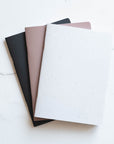 Three A5 Tom's Studio x Coffeenotes Notebooks in Black, Almond, and 'Grounds' (white), made from recycled coffee cups and eco-friendly materials.