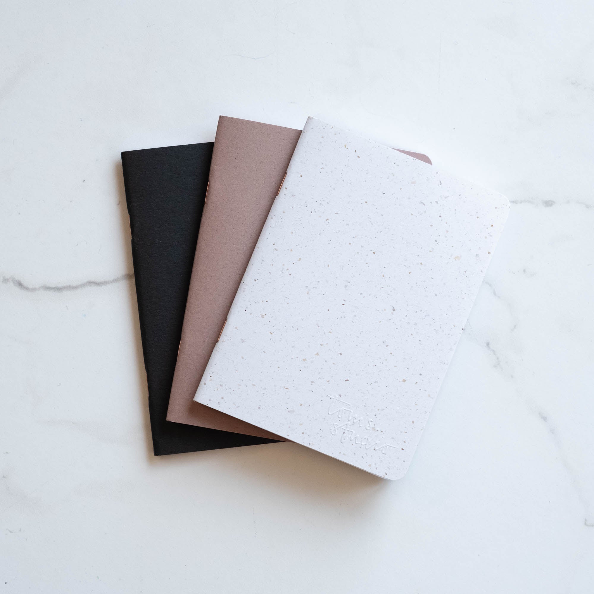 Three A6 Tom's Studio x Coffeenotes Notebooks in Black, Almond, and 'Grounds' (white), made from recycled coffee cups and eco-friendly materials.