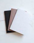 Three A6 Tom's Studio x Coffeenotes Notebooks in Black, Almond, and 'Grounds' (white), made from recycled coffee cups and eco-friendly materials.