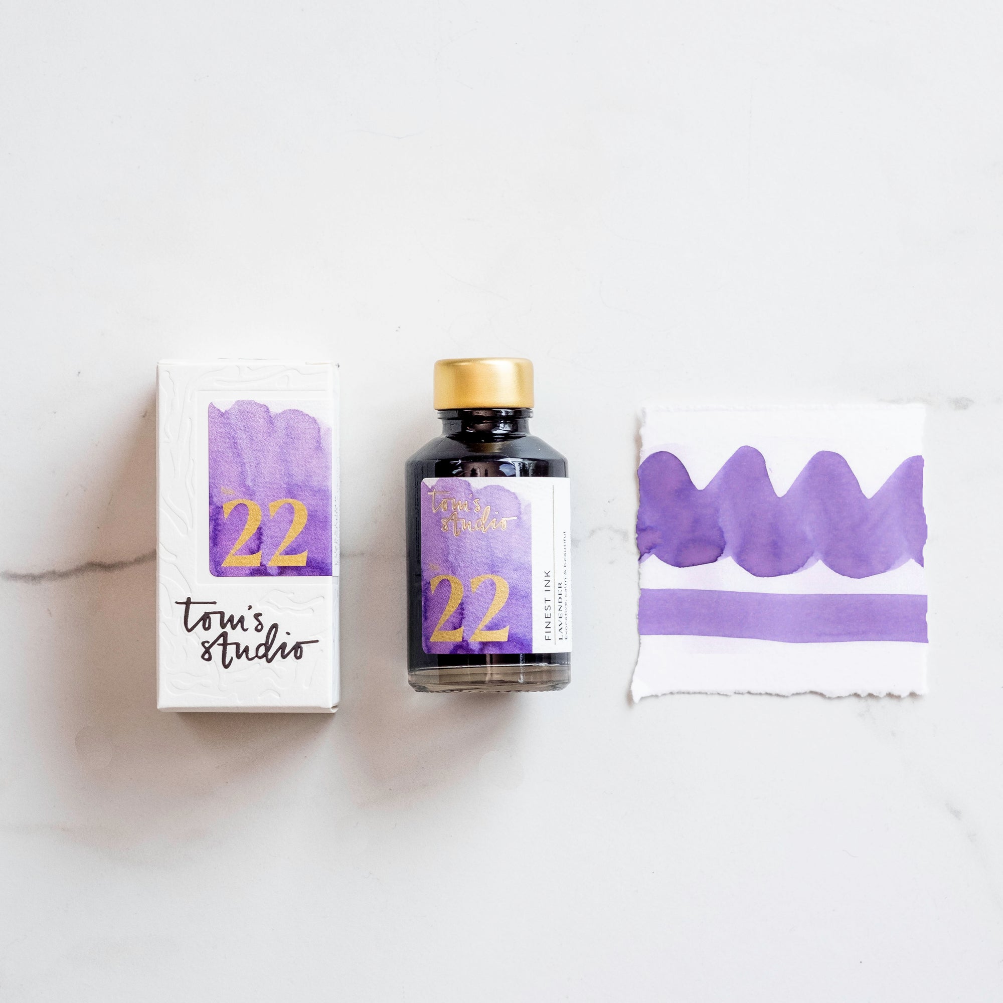 Tom's Studio Lavender Fountain Pen Ink – bottle with gold lid, boxed packaging featuring a purple and gold label marked '22', and a swatch of the vibrant lavender ink on textured paper.