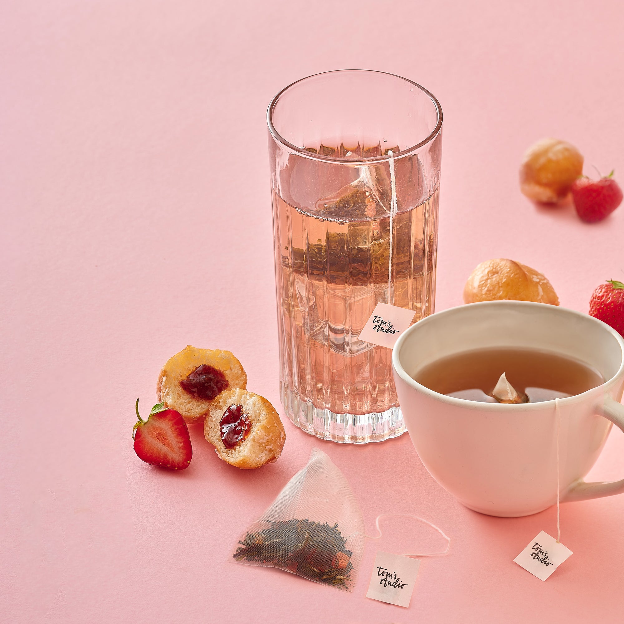 A tall glass and white teacup filled with pink-toned Jam Doughnut Tea sit on a pastel pink background, surrounded by a tea bag, fresh strawberries, and jam-filled doughnut halves. A tea bag with a Tom’s Studio tag is steeping in each drink.