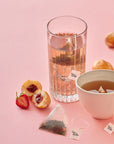 A tall glass and white teacup filled with pink-toned Jam Doughnut Tea sit on a pastel pink background, surrounded by a tea bag, fresh strawberries, and jam-filled doughnut halves. A tea bag with a Tom’s Studio tag is steeping in each drink.