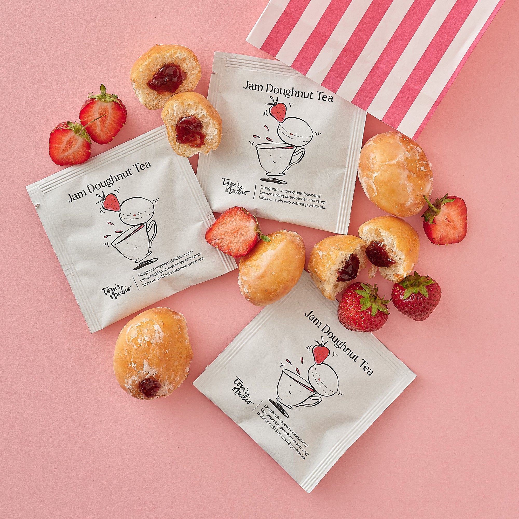 Several individually wrapped Jam Doughnut Tea sachets are styled with halved strawberries, whole and bitten jam doughnuts, and a pink-striped sweetshop bag. The packaging features a playful illustration of a jam doughnut bouncing into a teacup.