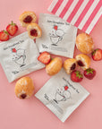 Several individually wrapped Jam Doughnut Tea sachets are styled with halved strawberries, whole and bitten jam doughnuts, and a pink-striped sweetshop bag. The packaging features a playful illustration of a jam doughnut bouncing into a teacup.