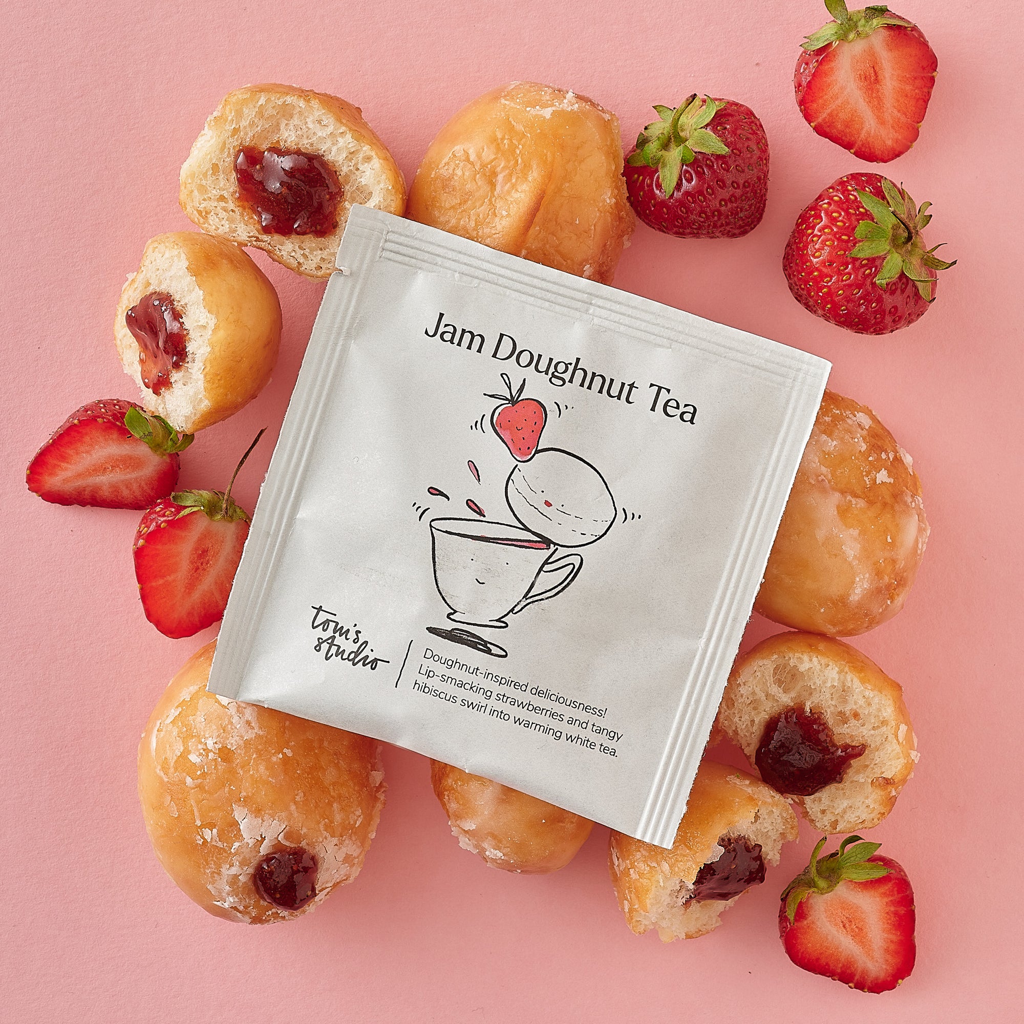 A single Jam Doughnut Tea sachet is centred on a pink background and surrounded by fresh strawberries and whole and halved jam doughnuts, showcasing the fruity and indulgent inspiration behind the blend.