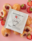 A single Jam Doughnut Tea sachet is centred on a pink background and surrounded by fresh strawberries and whole and halved jam doughnuts, showcasing the fruity and indulgent inspiration behind the blend.