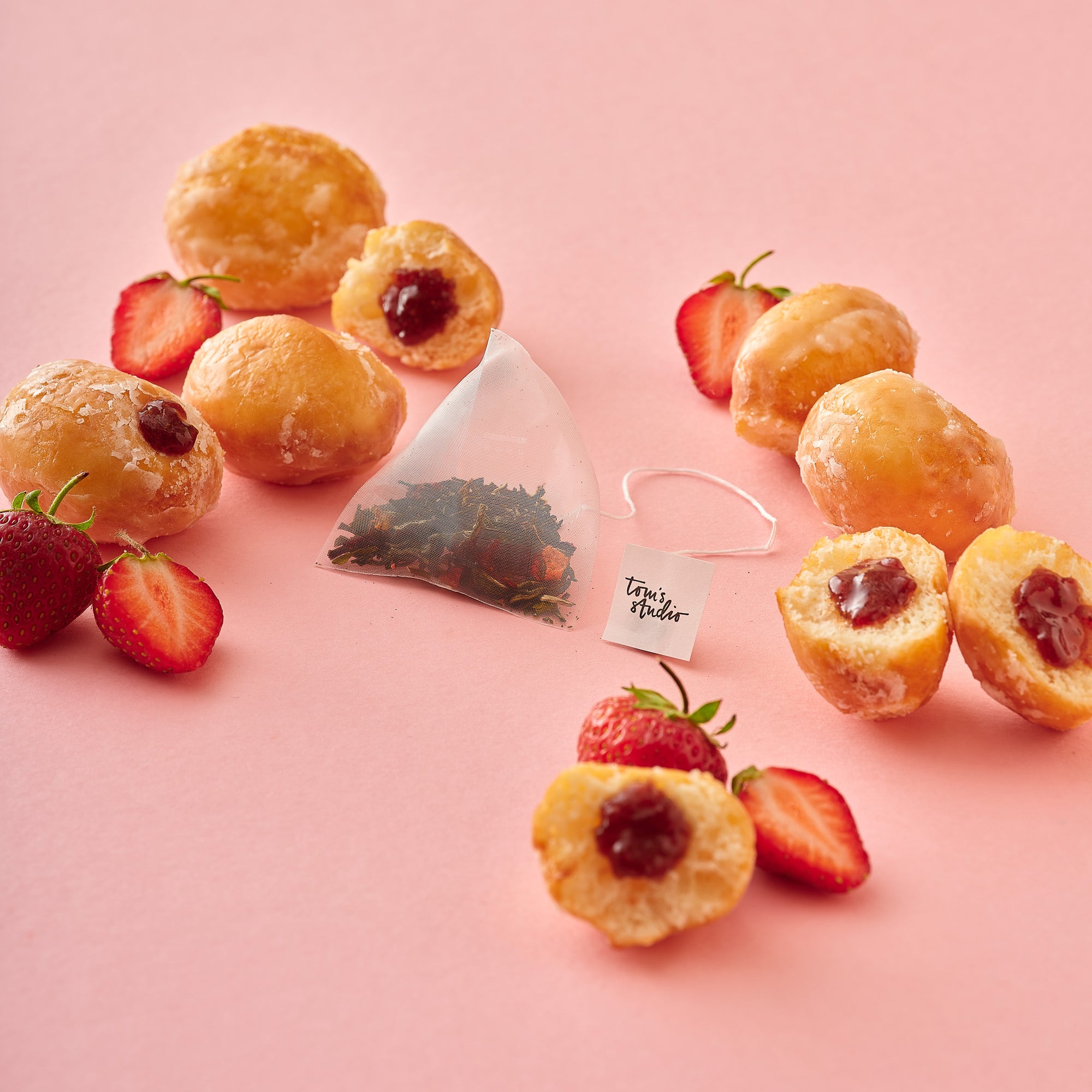 A pyramid-shaped Jam Doughnut Tea bag sits among scattered glazed jam doughnuts and sliced strawberries on a pastel pink surface. The bag reveals a colourful loose-leaf blend, highlighting the natural ingredients.