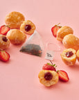 A pyramid-shaped Jam Doughnut Tea bag sits among scattered glazed jam doughnuts and sliced strawberries on a pastel pink surface. The bag reveals a colourful loose-leaf blend, highlighting the natural ingredients.