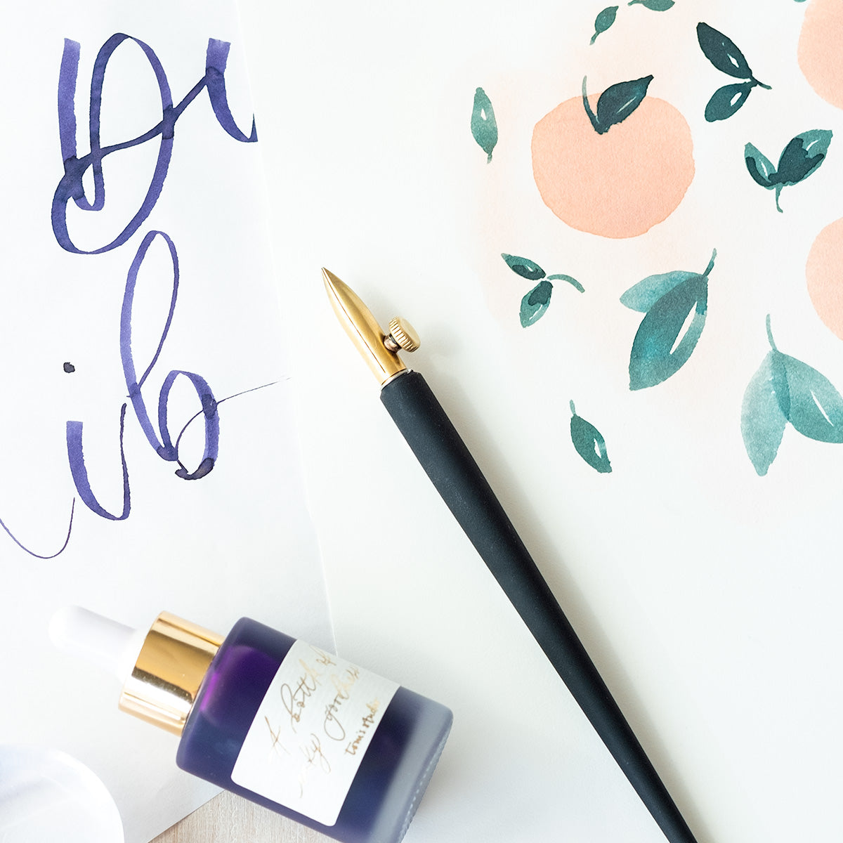 The One-Dip-Nib in Brass, shown fitted in the Original Flourish Straight Calligraphy Pen with a bottle of calligraphy ink on top of sample illustrations and lettering