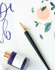 The One-Dip-Nib in Brass, shown fitted in the Original Flourish Straight Calligraphy Pen with a bottle of calligraphy ink on top of sample illustrations and lettering