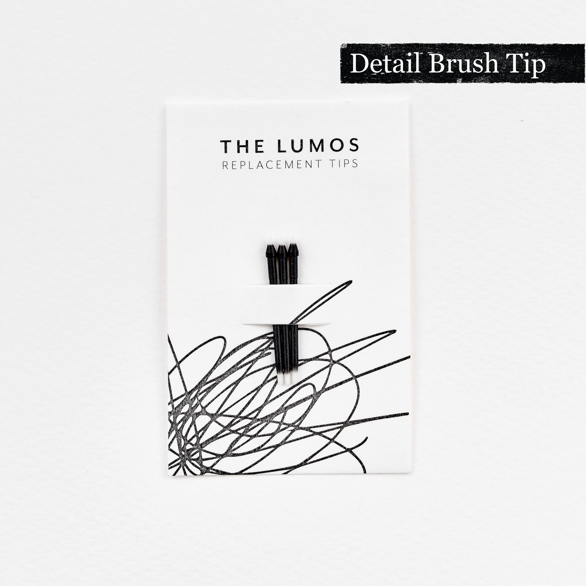 A pack of three Lumos replacement Detail Brush tips attached to a white backing card. The card features 'THE LUMOS REPLACEMENT TIPS' in uppercase black letters at the top, with an abstract black ink scribble design in the lower half. A black textured label in the upper right corner displays 'Detail Brush Tip' in white text.