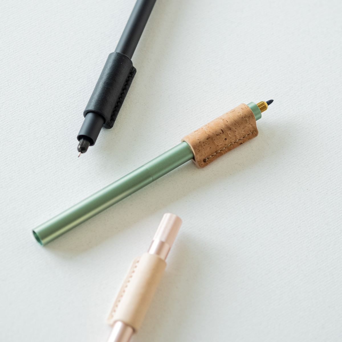 Handmade by Edward De Lacy, a pen grip and rollstop for the Lumos refillable pen – shown in black leather on the black Lumos, cork on the sage Lumos and natural leather on the rose gold Lumos