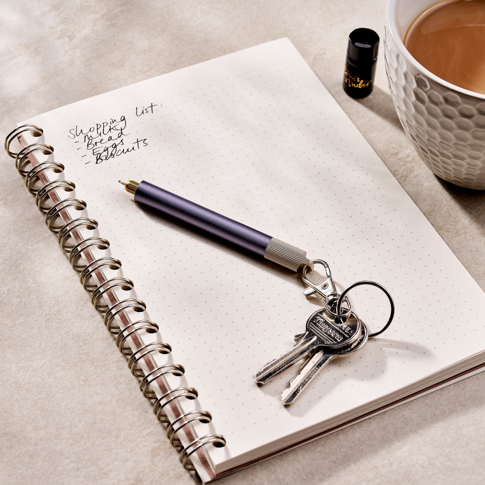 A Lumos Mini pen with a Loop Lid is attached to a set of keys and resting on an open notebook with handwritten notes. A cup of tea and a small ink bottle are in the background, creating a warm and creative workspace setting.