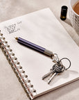 A Lumos Mini pen with a Loop Lid is attached to a set of keys and resting on an open notebook with handwritten notes. A cup of tea and a small ink bottle are in the background, creating a warm and creative workspace setting.