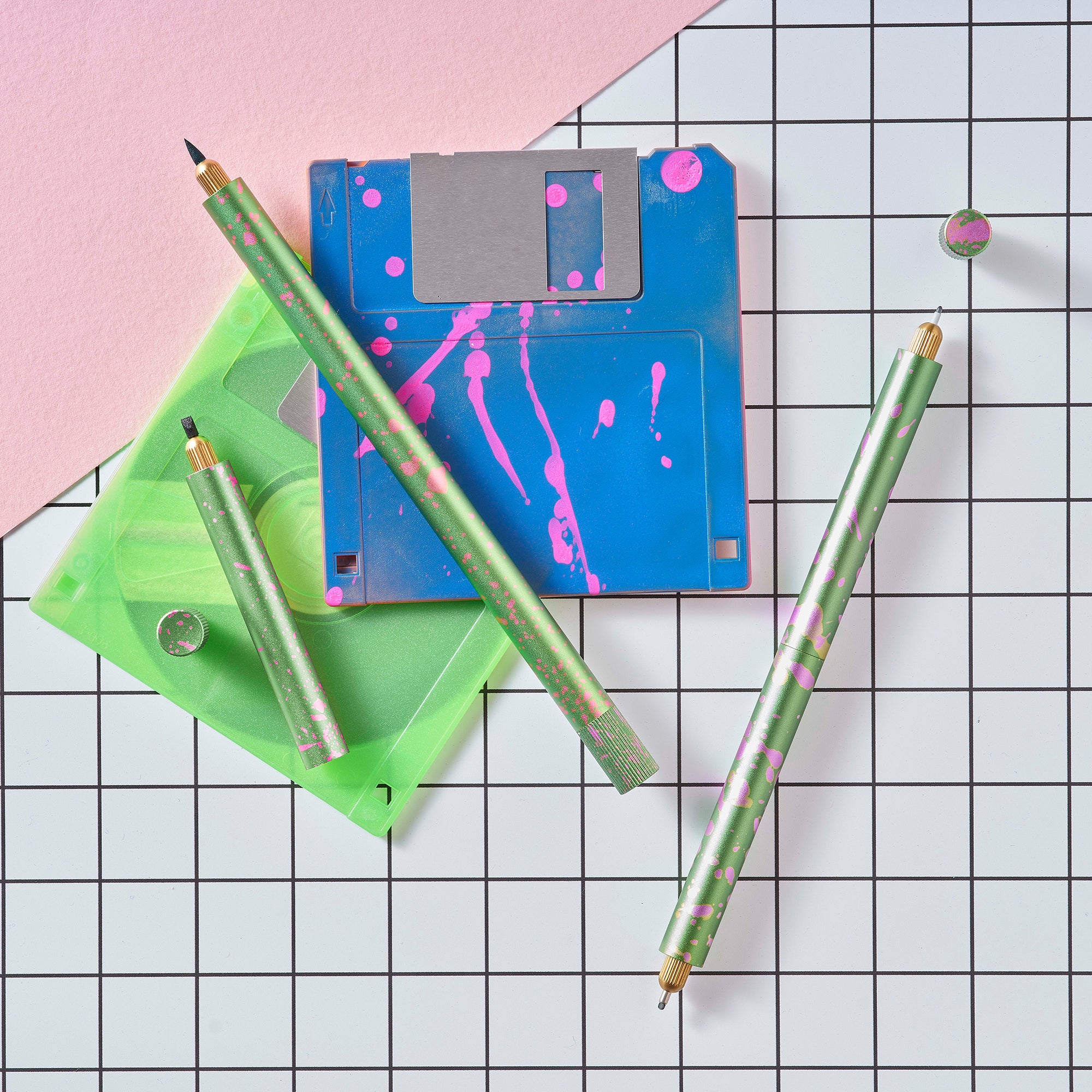Close-up of Lumos Splash pens in Appletini green with pink splatter, styled with blue and green floppy disks on a grid background.