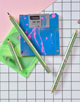 Close-up of Lumos Splash pens in Appletini green with pink splatter, styled with blue and green floppy disks on a grid background.