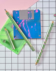Close-up of Lumos Splash pens in Appletini green with pink splatter, styled with blue and green floppy disks on a grid background.
