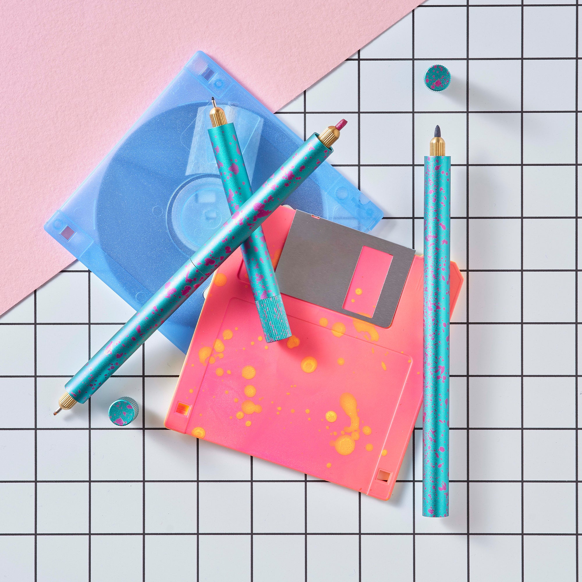 Close-up of Lumos Splash pens in Blue Raspberry colourway with pink splatter, styled with neon pink and blue floppy disks on a grid background.