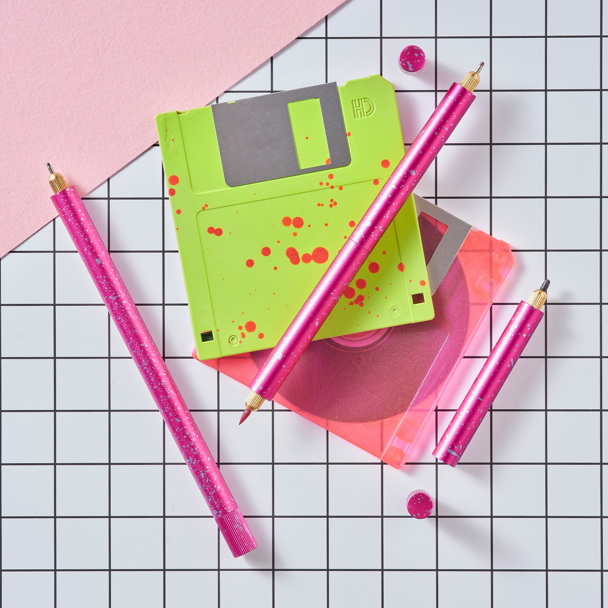 Close-up of Lumos Splash pens in Bubblegum pink with blue splatter, styled with neon green and pink floppy disks on a grid background.
