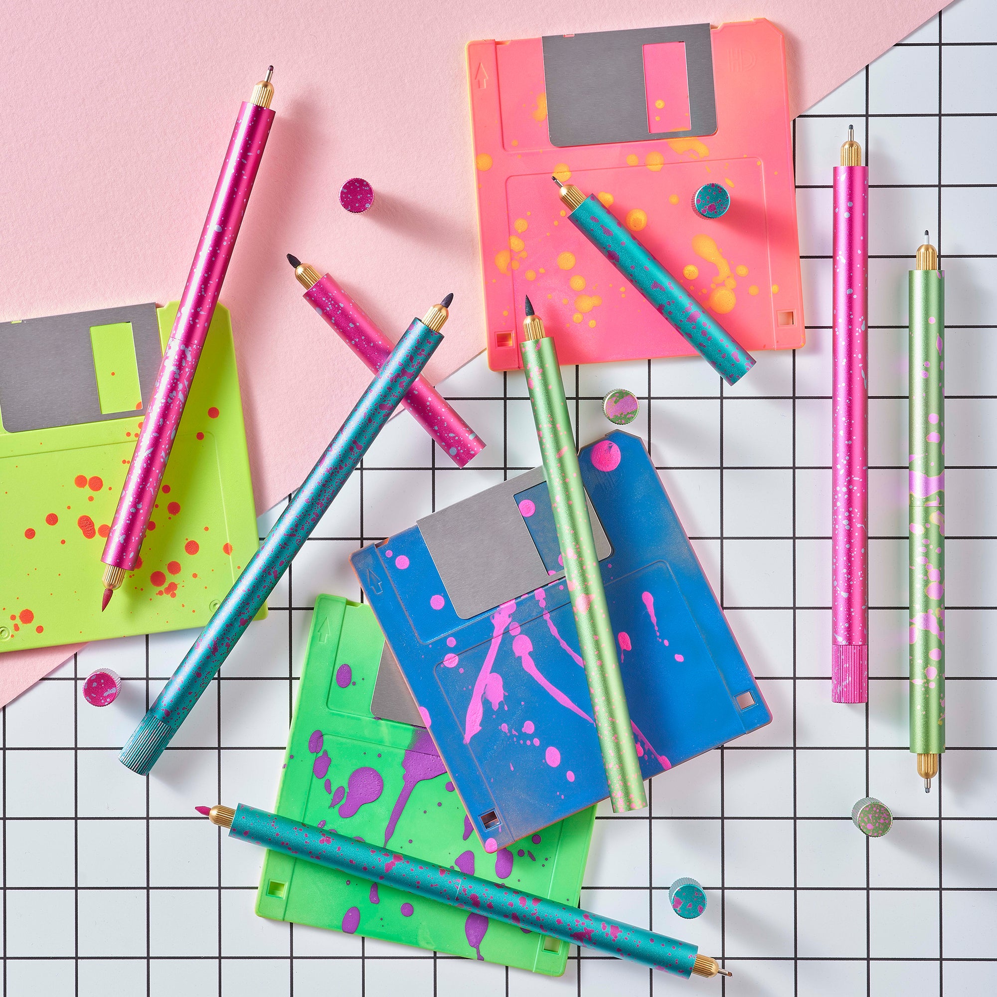 Flat lay of Lumos Splash pens in all three neon colourways with paint-splatter patterns, arranged with bright floppy disks on a grid background.