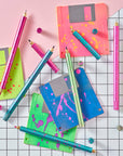 Flat lay of Lumos Splash pens in all three neon colourways with paint-splatter patterns, arranged with bright floppy disks on a grid background.