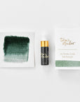 A mini ink sample bottle of Ivy green fountain pen ink by Tom’s Studio, displayed alongside a swatch of the ink on textured paper and its matching packaging. The bottle is small, with a gold cap and a white pipette dropper, and features 'Tom’s Studio' in gold lettering. The ink swatch shows a rich, shimmering hue with a gradient from deep to lighter tones. The packaging, a small white card, has 'Tom’s Studio' embossed in gold at the top, with a brushstroke of the ink colour at the bottom.