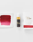 A mini ink sample bottle of Cassis red fountain pen ink by Tom’s Studio, displayed alongside a swatch of the ink on textured paper and its matching packaging. The bottle is small, with a gold cap and a white pipette dropper, and features 'Tom’s Studio' in gold lettering. The ink swatch shows a rich, shimmering hue with a gradient from deep to lighter tones. The packaging, a small white card, has 'Tom’s Studio' embossed in gold at the top, with a brushstroke of the ink colour at the bottom.