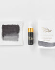 A mini ink sample bottle of Dove Grey fountain pen ink by Tom’s Studio, displayed alongside a swatch of the ink on textured paper and its matching packaging. The bottle is small, with a gold cap and a white pipette dropper, and features 'Tom’s Studio' in gold lettering. The ink swatch shows a rich, shimmering hue with a gradient from deep to lighter tones. The packaging, a small white card, has 'Tom’s Studio' embossed in gold at the top, with a brushstroke of the ink colour at the bottom.