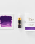 A mini ink sample bottle of Iris purple fountain pen ink by Tom’s Studio, displayed alongside a swatch of the ink on textured paper and its matching packaging. The bottle is small, with a gold cap and a white pipette dropper, and features 'Tom’s Studio' in gold lettering. The ink swatch shows a rich, shimmering hue with a gradient from deep to lighter tones. The packaging, a small white card, has 'Tom’s Studio' embossed in gold at the top, with a brushstroke of the ink colour at the bottom.