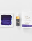 A mini ink sample bottle of Juniper purple fountain pen ink by Tom’s Studio, displayed alongside a swatch of the ink on textured paper and its matching packaging. The bottle is small, with a gold cap and a white pipette dropper, and features 'Tom’s Studio' in gold lettering. The ink swatch shows a rich, shimmering hue with a gradient from deep to lighter tones. The packaging, a small white card, has 'Tom’s Studio' embossed in gold at the top, with a brushstroke of the ink colour at the bottom.
