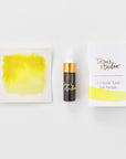 A mini ink sample bottle of Lemon Yellow fountain pen ink by Tom’s Studio, displayed alongside a swatch of the ink on textured paper and its matching packaging. The bottle is small, with a gold cap and a white pipette dropper, and features 'Tom’s Studio' in gold lettering. The ink swatch shows a rich, shimmering hue with a gradient from deep to lighter tones. The packaging, a small white card, has 'Tom’s Studio' embossed in gold at the top, with a brushstroke of the ink colour at the bottom.