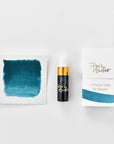A mini ink sample bottle of Loch Ness blue fountain pen ink by Tom’s Studio, displayed alongside a swatch of the ink on textured paper and its matching packaging. The bottle is small, with a gold cap and a white pipette dropper, and features 'Tom’s Studio' in gold lettering. The ink swatch shows a rich, shimmering hue with a gradient from deep to lighter tones. The packaging, a small white card, has 'Tom’s Studio' embossed in gold at the top, with a brushstroke of the ink colour at the bottom.