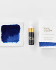 A mini ink sample bottle of Marianas blue fountain pen ink by Tom’s Studio, displayed alongside a swatch of the ink on textured paper and its matching packaging. The bottle is small, with a gold cap and a white pipette dropper, and features 'Tom’s Studio' in gold lettering. The ink swatch shows a rich, shimmering hue with a gradient from deep to lighter tones. The packaging, a small white card, has 'Tom’s Studio' embossed in gold at the top, with a brushstroke of the ink colour at the bottom.
