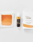 A mini ink sample bottle of Marmalade orange fountain pen ink by Tom’s Studio, displayed alongside a swatch of the ink on textured paper and its matching packaging. The bottle is small, with a gold cap and a white pipette dropper, and features 'Tom’s Studio' in gold lettering. The ink swatch shows a rich, shimmering hue with a gradient from deep to lighter tones. The packaging, a small white card, has 'Tom’s Studio' embossed in gold at the top, with a brushstroke of the ink colour at the bottom.