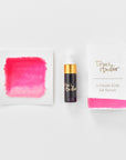 A mini ink sample bottle of Rambling Rose pink fountain pen ink by Tom’s Studio, displayed alongside a swatch of the ink on textured paper and its matching packaging. The bottle is small, with a gold cap and a white pipette dropper, and features 'Tom’s Studio' in gold lettering. The ink swatch shows a rich, shimmering hue with a gradient from deep to lighter tones. The packaging, a small white card, has 'Tom’s Studio' embossed in gold at the top, with a brushstroke of the ink colour at the bottom.