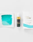 A mini ink sample bottle of Sunny Teal blue fountain pen ink by Tom’s Studio, displayed alongside a swatch of the ink on textured paper and its matching packaging. The bottle is small, with a gold cap and a white pipette dropper, and features 'Tom’s Studio' in gold lettering. The ink swatch shows a rich, shimmering hue with a gradient from deep to lighter tones. The packaging, a small white card, has 'Tom’s Studio' embossed in gold at the top, with a brushstroke of the ink colour at the bottom.